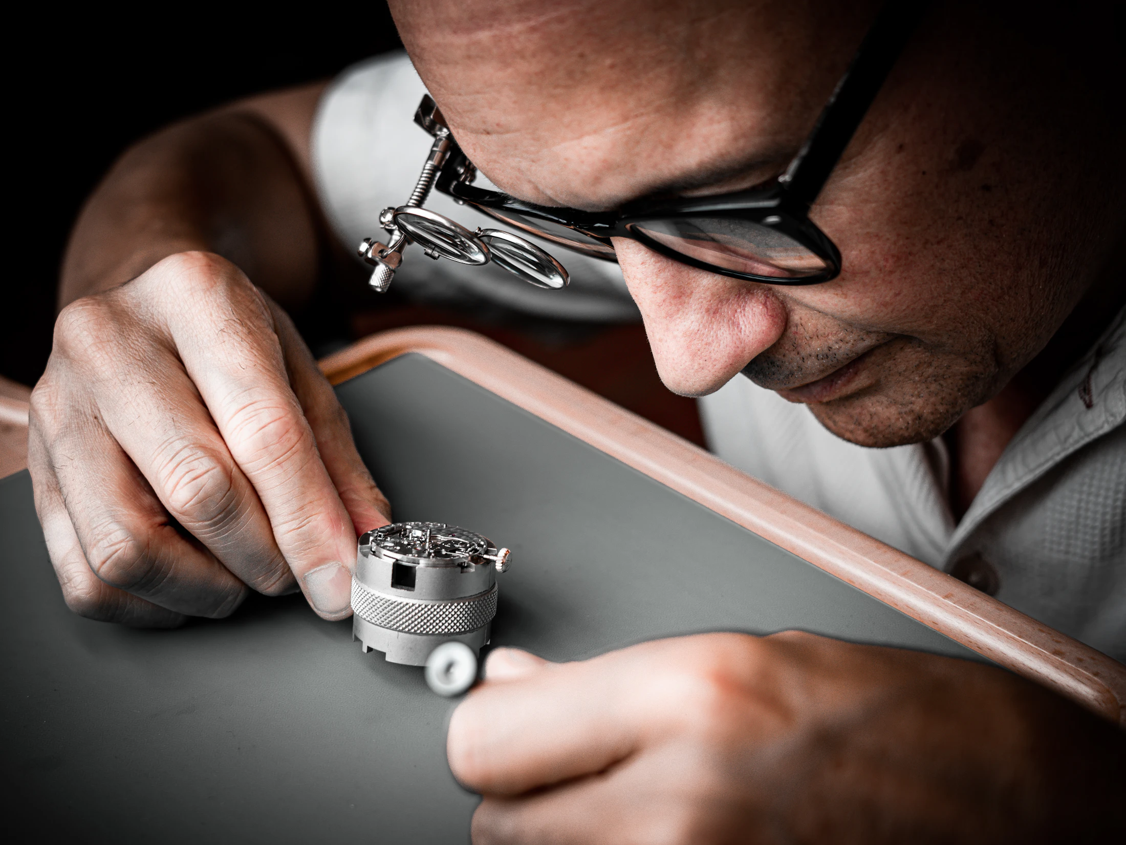 A watchmaker bent low over a movement in its holder, a double loupe clipped to his glasses.