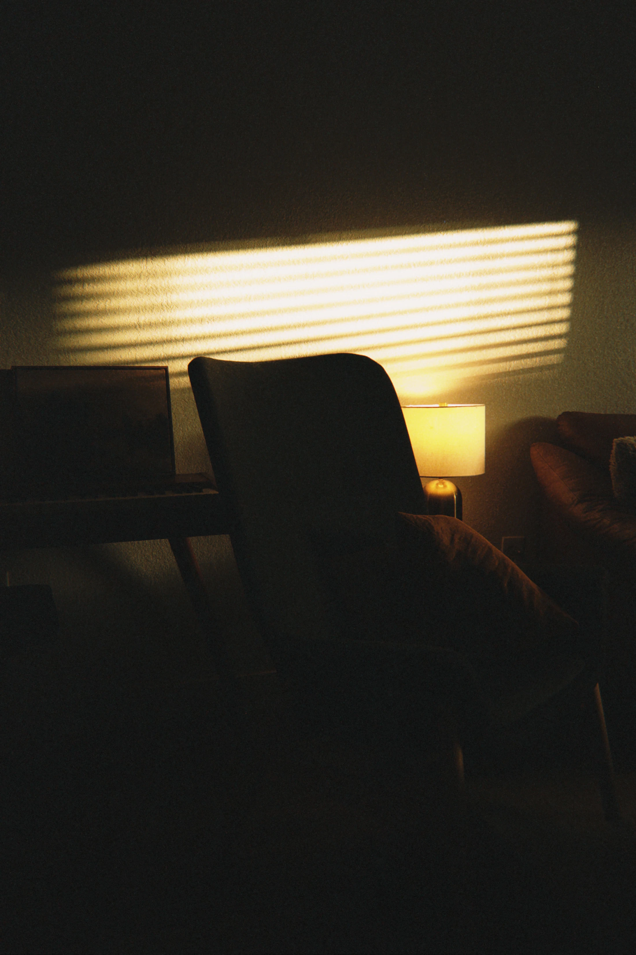 A chair and a low lamp in a dark room, late sun striping the wall.